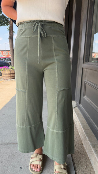 A person wearing olive green wide-leg drawstring pants with contrast stitching, paired with a cream ribbed top and beige sandals, standing on a sidewalk.