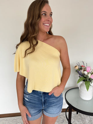 Woman wearing a yellow one-shoulder top and denim shorts, standing indoors.