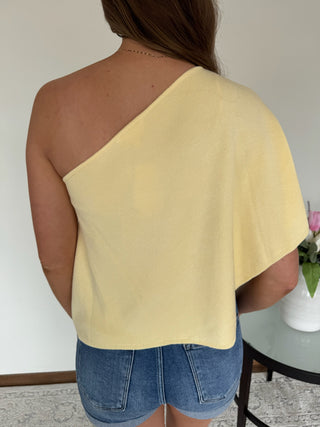 Person wearing a yellow one-shoulder top and denim shorts against a neutral background