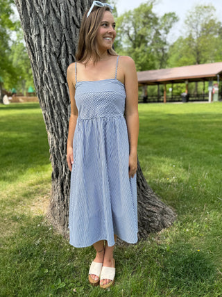 Woman in a blue striped dress standing next to a tree in a park.