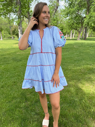 Woman in a light blue dress with red trim standing in a park.
