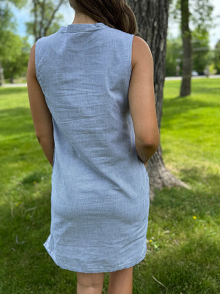 Person wearing a light blue sleeveless dress standing in a grassy area with trees.