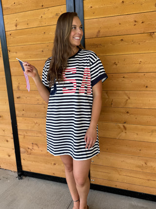 Woman wearing a black and white striped dress with red text, standing against a wooden wall.