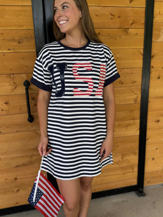Woman wearing a striped dress with a large letter 'S' holding an American flag against a wooden wall.