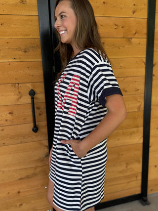 Woman wearing a black and white striped dress with red text, standing against a wooden wall.