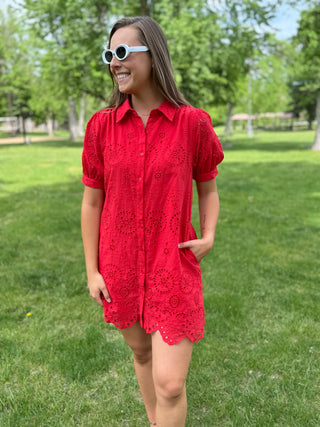 Woman wearing a red dress and white sunglasses standing in a grassy area with trees in the background