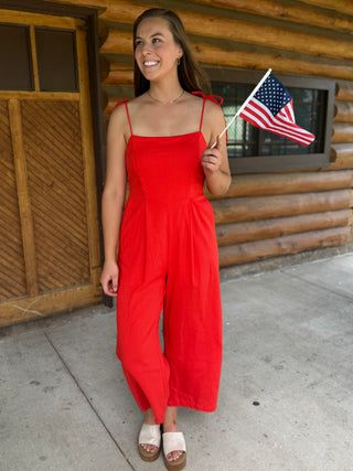 Woman in a red jumpsuit holding an American flag in front of a wooden building.