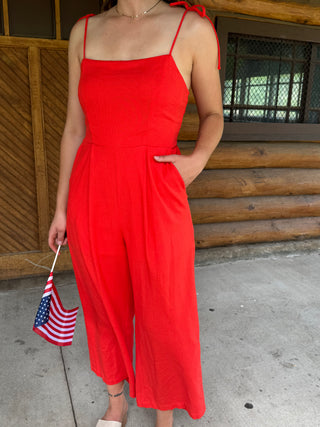 Person wearing a red jumpsuit holding an American flag in front of a wooden building.