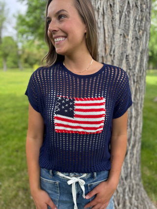 Woman wearing a navy blue sweater with an American flag design, standing outdoors near a tree.