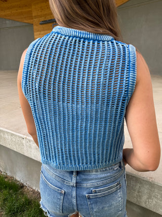 Person wearing a blue knitted vest and jeans outdoors.