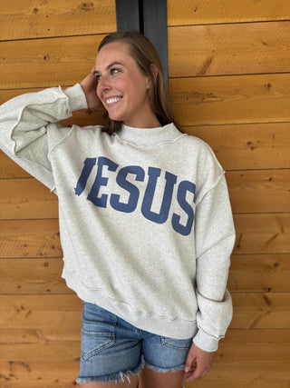 Person wearing a sweatshirt with 'JESUS' printed on it, standing against a wooden wall.