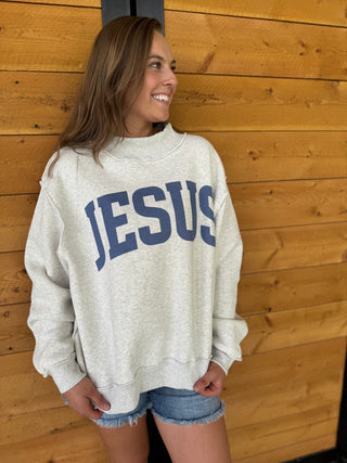 Woman wearing a sweatshirt with 'JESUS' printed on it against a wooden wall.