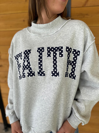 Person wearing a gray sweatshirt with 'FAITH' printed in black polka dots.
