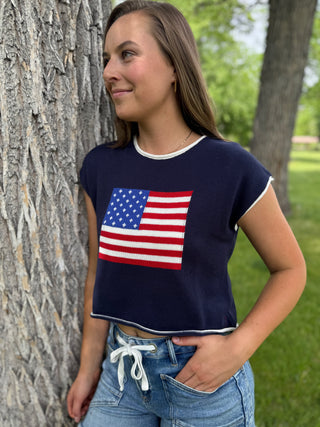 Woman wearing a navy blue t-shirt with an American flag design, standing outdoors near trees.