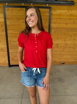 Woman wearing a red checkered blouse and denim shorts standing in front of a wooden wall.