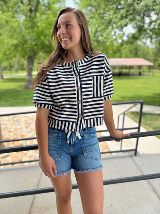 Woman wearing a striped shirt and denim shorts standing outdoors with trees and a gazebo in the background.