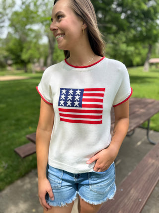 Woman wearing a white sweater with an American flag design outdoors.