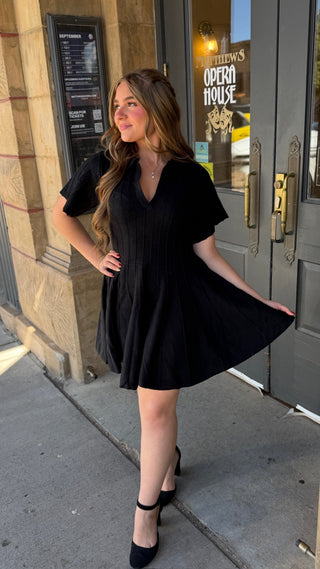 A woman wearing a black short-sleeve pleated mini dress and black heels stands on a city sidewalk in front of a building entrance with gray doors and a sign reading 'Opera House.'.