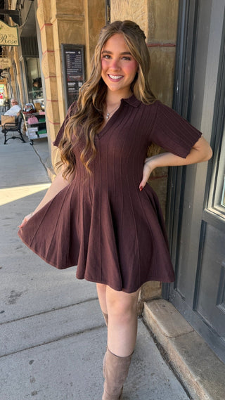 A woman wearing a short-sleeve, collared, flared mini dress in a deep plum-brown color, standing outdoors on a city sidewalk next to a building.
