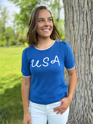 Woman wearing a blue shirt with 'USA' text, standing outdoors next to a tree.