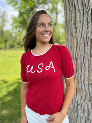 Woman wearing a red 'USA' shirt standing next to a tree in a park.