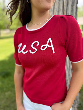 Person wearing a red sweater with 'USA' printed on it, standing outdoors.