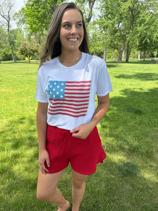 Woman wearing a white t-shirt with an American flag design and red shorts in a park.