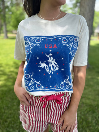 Person wearing a white t-shirt with a blue graphic design featuring 'USA' and a rodeo rider, standing outdoors.