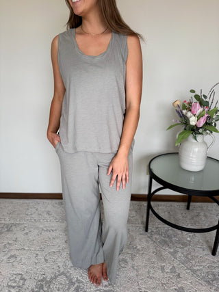 Woman wearing a gray sleeveless top and pants standing in a room with a table and flowers.