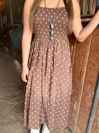 Woman wearing a brown dress with white polka dots standing against a wooden wall.