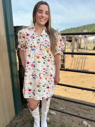 Woman in a floral dress standing in front of a fence with horses in the background
