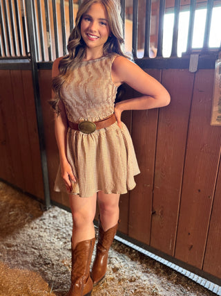 Woman in a checkered dress with a brown belt and boots standing in a stable.