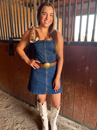 Woman in a blue denim dress with a brown belt and white cowboy boots standing in a barn.