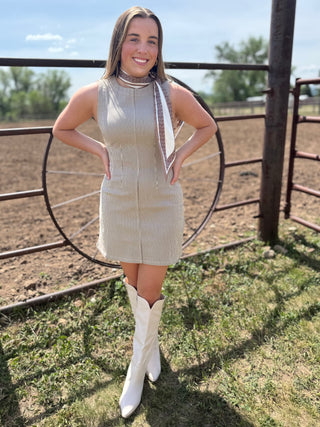 Woman in a beige dress and white boots standing in front of a metal gate with a rural background.