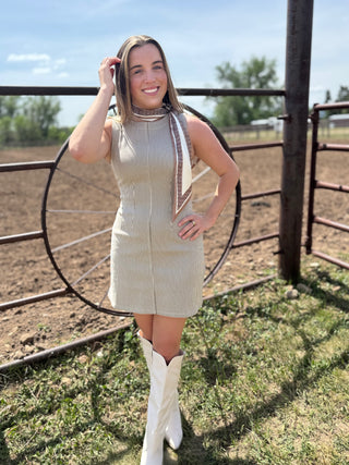 Woman in a beige dress and white boots standing in a rural setting with a fence and open field.