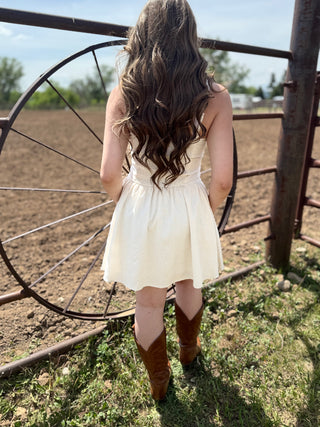 Person in a white dress and brown boots standing near a rustic metal gate with a plowed field in the background.