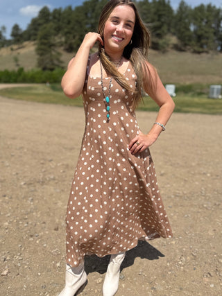 Woman in a brown polka dot dress standing outdoors with trees in the background