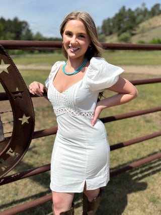 Woman in a white dress standing in front of a rustic metal gate with a scenic background.