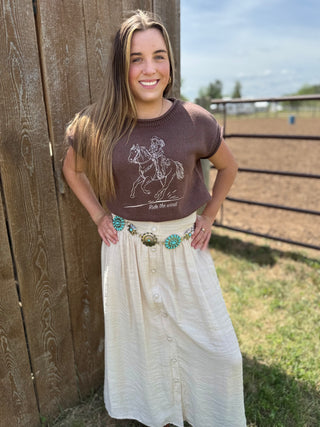Woman wearing a brown t-shirt with a graphic design and a white skirt standing next to a wooden fence.