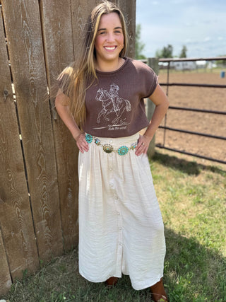 Woman standing outdoors near a wooden fence, wearing a brown graphic t-shirt and white skirt.