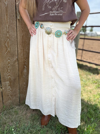 Person wearing a long white skirt with decorative buttons and a belt with turquoise stones, standing against a wooden fence.