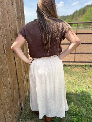 Person wearing a brown top and white skirt standing against a wooden fence with a scenic background.
