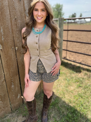 Woman standing outdoors near a wooden fence wearing a beige vest, patterned shorts, and brown boots.