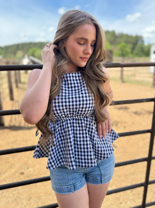 Woman wearing a black and white checkered top and denim shorts standing in front of a fence with a rural background.