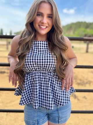 Woman wearing a black and white checkered top and blue shorts standing in front of a fence with a scenic background.