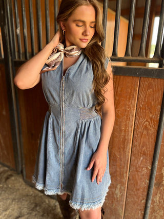 Woman in a denim dress standing in front of wooden stalls, holding a patterned scarf.