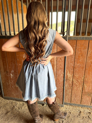 Woman in a denim dress and brown boots standing in front of wooden stable doors.