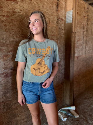 Woman wearing a 'Cowboy Country' t-shirt with a guitar and cowboy hat graphic, standing in a rustic setting.