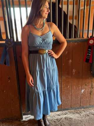 Woman in a blue dress standing in an equestrian setting with horse stalls in the background.
