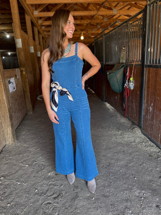 Woman wearing a blue denim jumpsuit in an indoor setting with wooden beams and a horse stall.